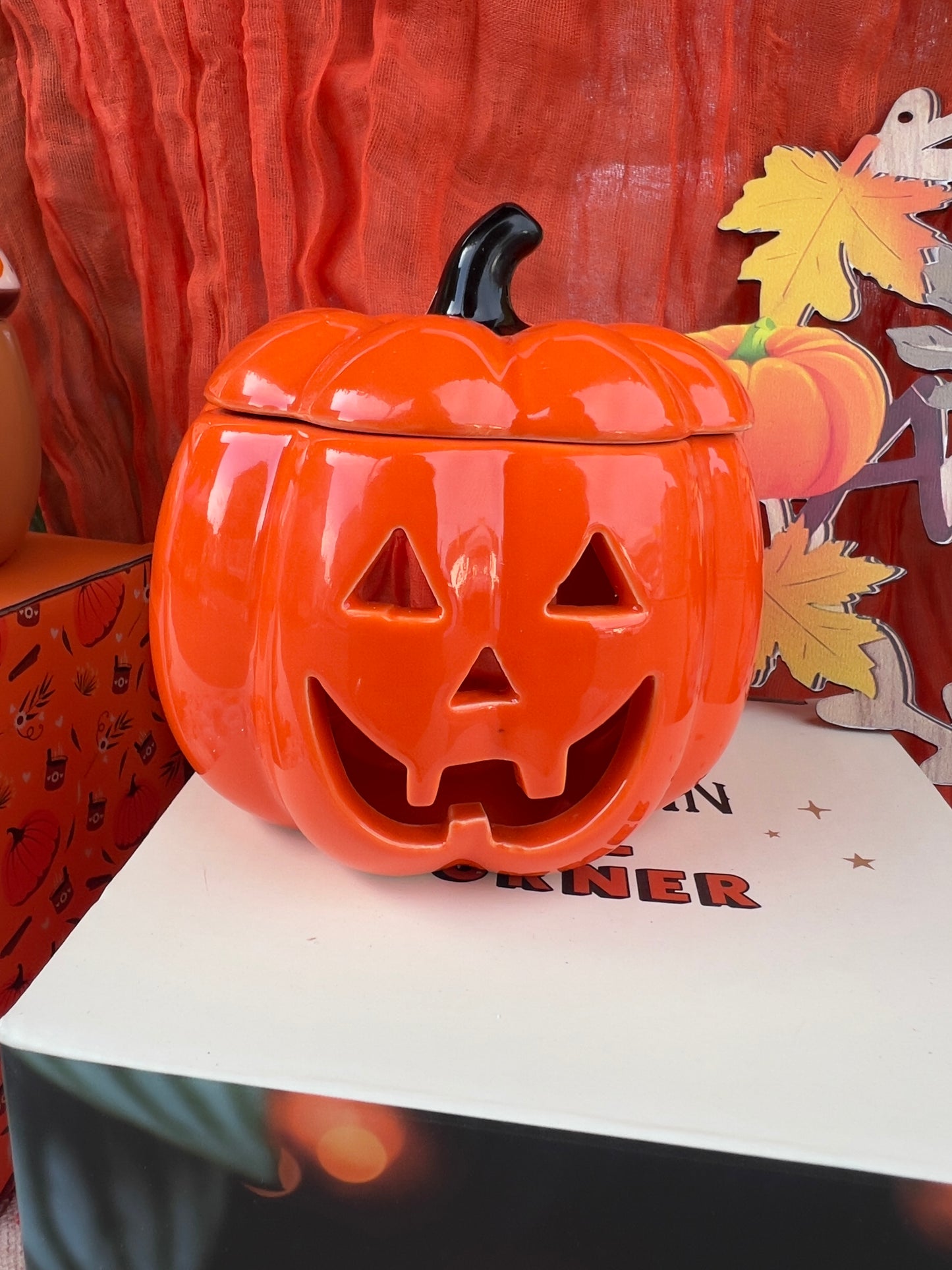 Orange pumpkin-shaped wax melt/oil burner with a carved face on a Halloween-themed background.